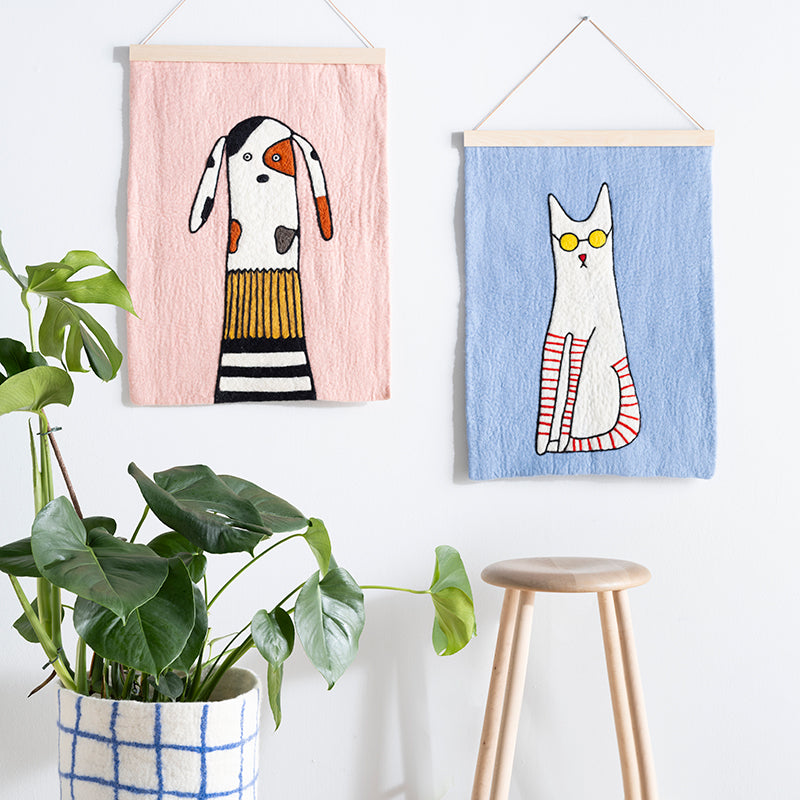 Two wall hangings of a dog and a cat on a white wall with a plant and stool in the foreground.