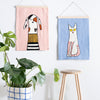 Two wall hangings of a dog and a cat on a white wall with a plant and stool in the foreground.