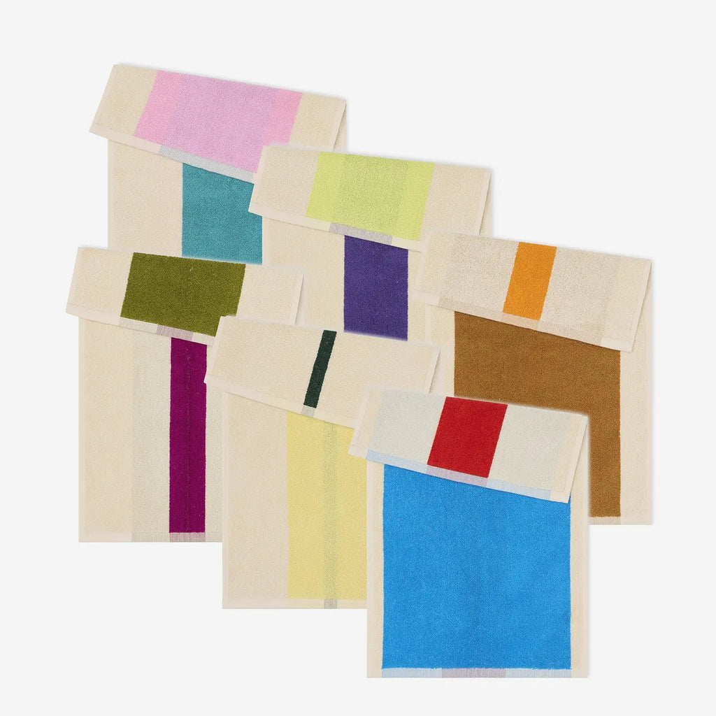 A set of folded cream-colored towels with large, solid color blocks in pink, teal, olive green, purple, yellow, brown, orange, magenta, and blue, arranged in a fan-like pattern on a light gray background.