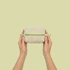 A person's hands holding a small rectangular beige linen pouch with a green band and white end-tags against a solid pale yellow-green background.