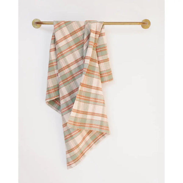 A plaid kitchen towel in muted sage green, terracotta orange, and cream hangs draped from a brass wall-mounted rod against a plain white wall.