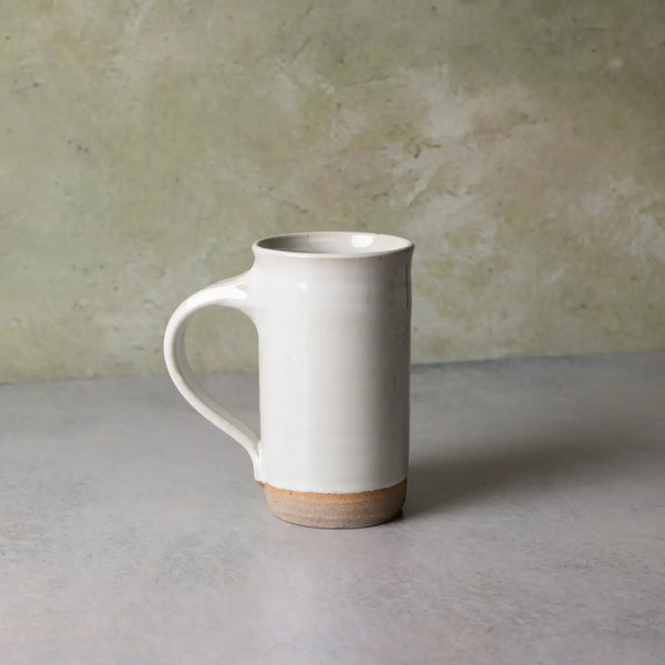 A tall, cylindrical ceramic mug with a curved handle, featuring a glossy off-white glaze and a natural, unglazed tan clay base, set against a textured light gray and beige background.