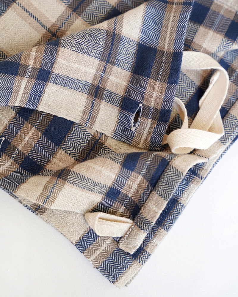 A close-up of a folded plaid kitchen cloth, featuring a navy blue, beige, and brown herringbone pattern with cream canvas straps on a white background.
