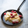 Two folded crepes on a frying pan with raspberries on a light gray background