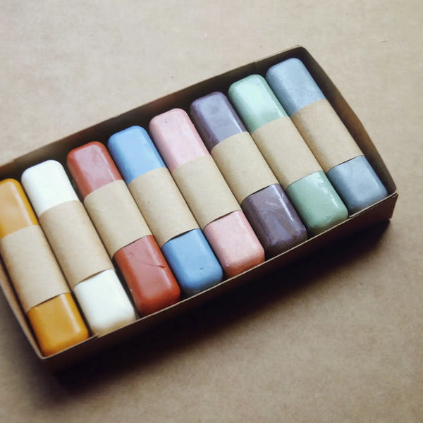 A set of pastel-colored wax crayons in a cardboard box, each wrapped with a paper band. The crayons come in various colors including yellow, white, terracotta, light blue, pink, purple, mint green, and light blue.