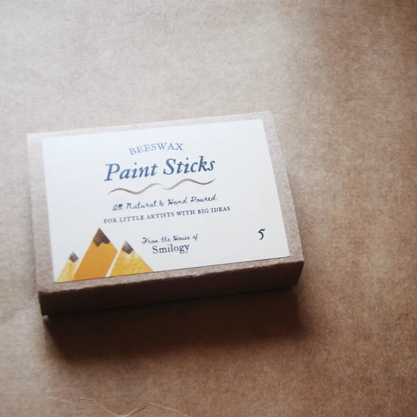 A box of Beeswax Paint Sticks by Smiley on a textured brown paper background. The packaging features a white label with blue text and yellow pencil illustrations.