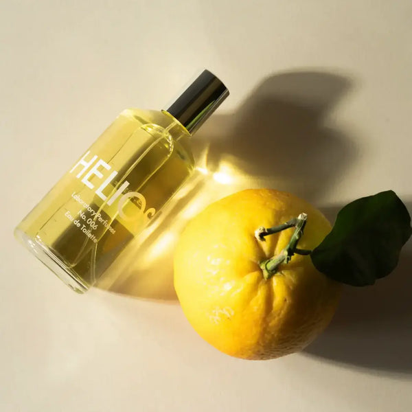 Perfume bottle labeled 'HELIOS' next to a lemon on a light background