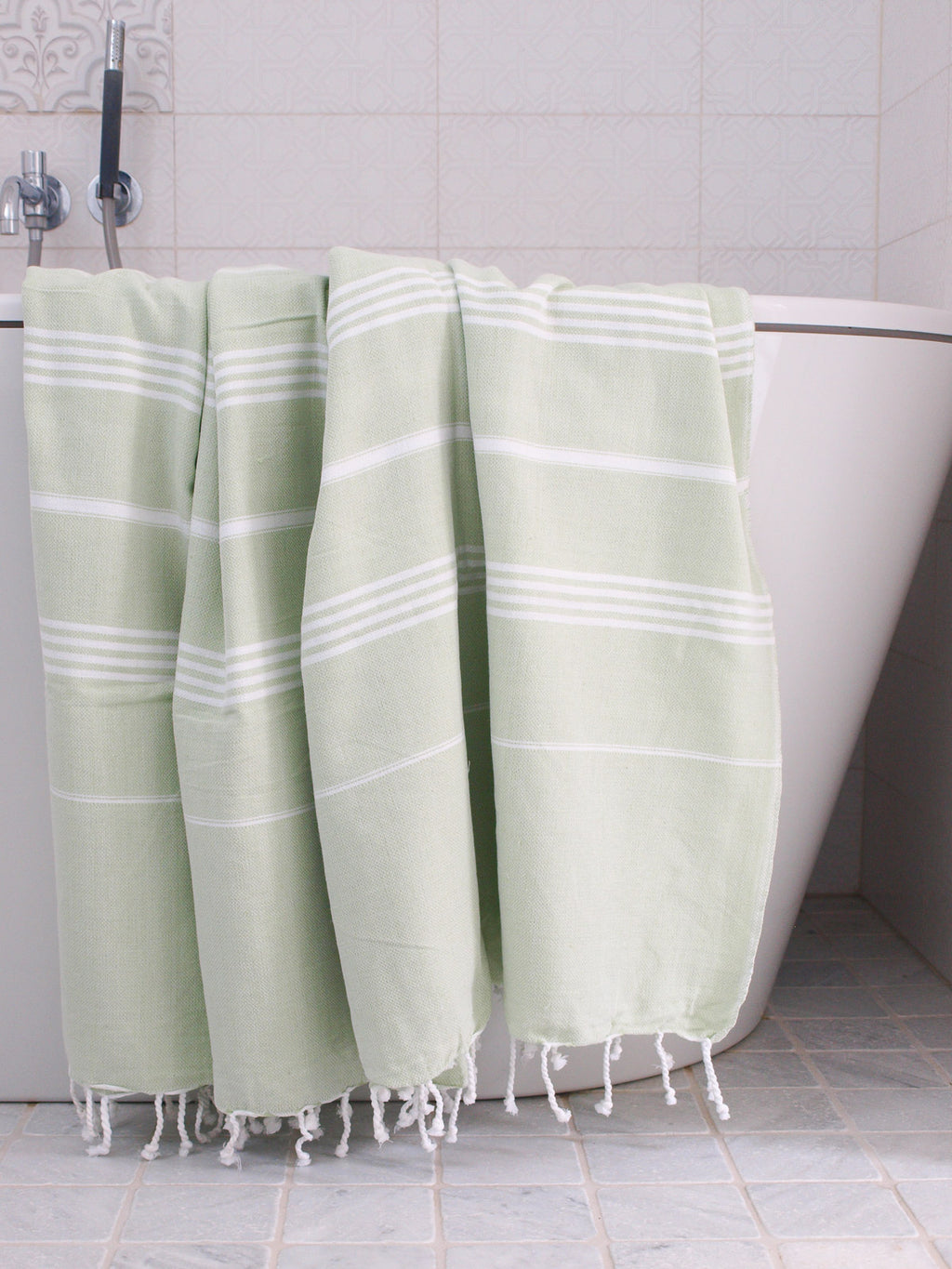 light green towel with white stripes and tassels hanging on a bath tub in a bathroom.