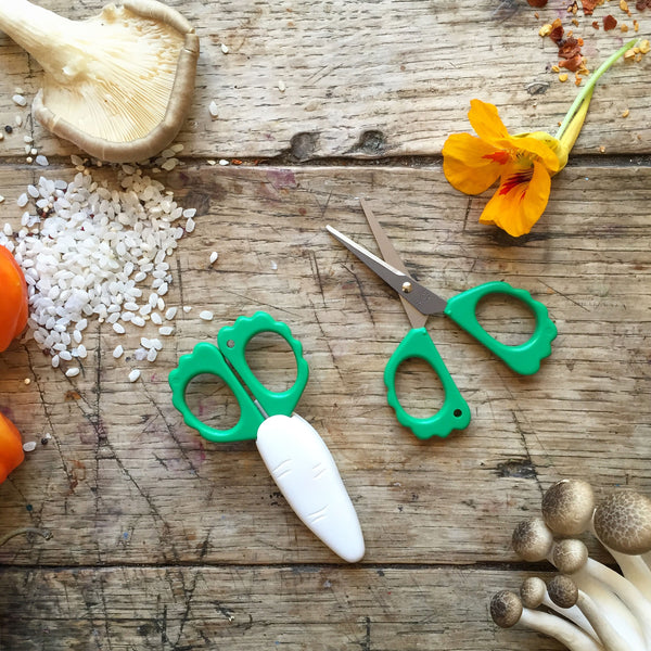 Pair of green-handled scissors on a wooden surface with vegetables and a flower.