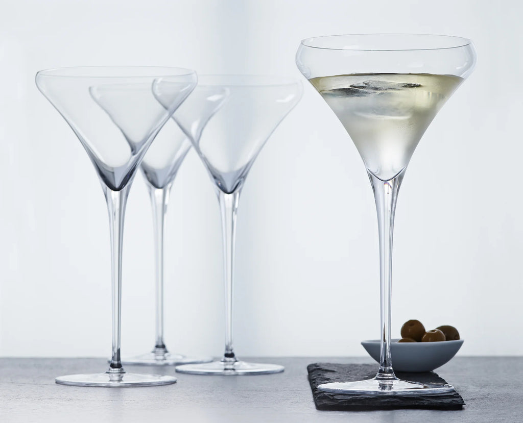 Four clear martini glasses on a gray surface against a white background, with one glass filled with a pale drink and a small dish of olives beside it.