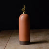 A tall, matte terracotta-colored ceramic oil bottle with a gold-tone metal spout and a dark brown base, set on a rustic wooden surface against a solid black background.