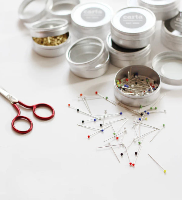 Scissors and pins on a white surface with small containers in the background.