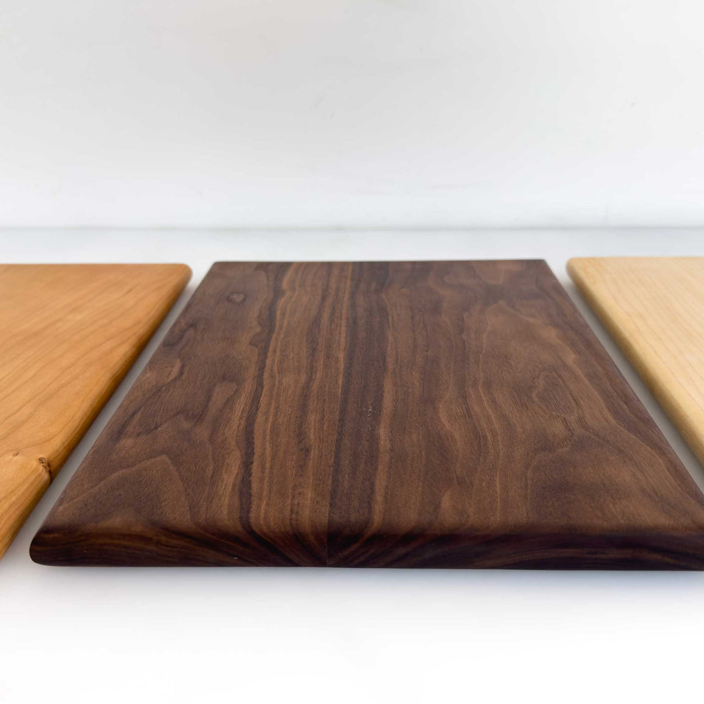 Three rectangular wooden cutting boards with rounded corners displayed side-by-side, featuring different wood grain patterns and tones: a dark brown walnut board in the center, flanked by two lighter natural wood boards on either side, set against a plain white background.