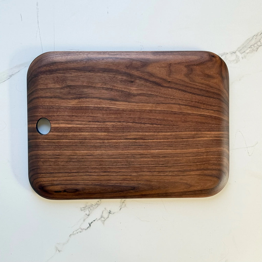 A rectangular wooden cutting board with rounded corners and a small circular hole on the left side, shown on a white marble surface.